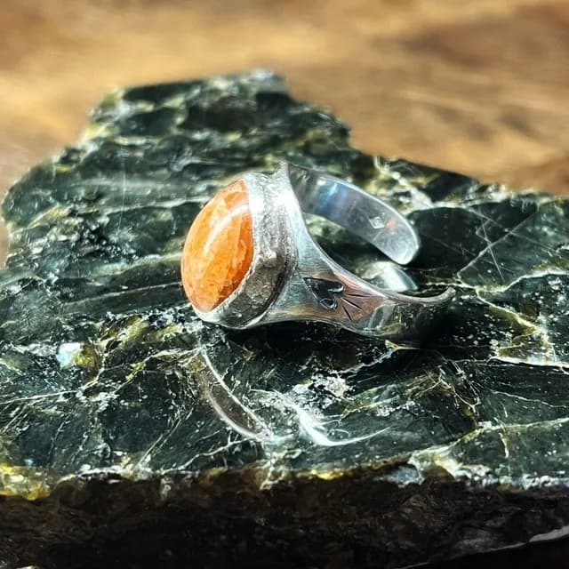 Bague en argent avec pierre de soleil orange et cœur gravé, posée sur une roche sombre.