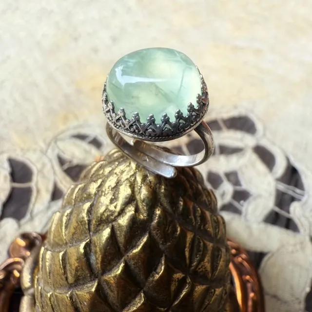 Bague en argent avec un béryl vert arrondi, posée sur un support doré en forme de pomme de pin.