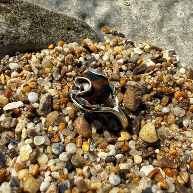Bague en argent et jaspe océan vert et marron, posée de trois-quarts sur un lit de petits galets colorés.