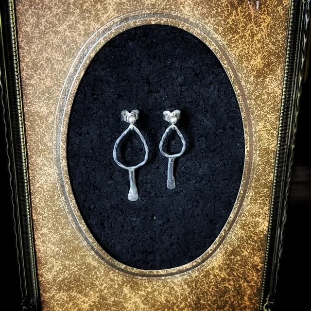 Boucles d'oreilles argentées de forme ovale allongée, présentées dans un cadre doré ancien au fond noir.