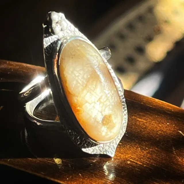 Bague en métal argenté avec une grande pierre ovale beige, posée sur un support en bois sombre.