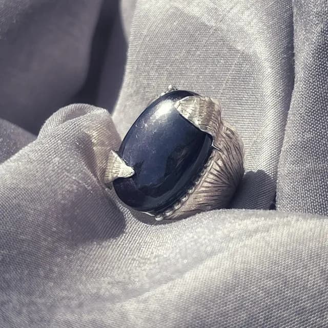 Bague en argent gravé sertie d'une pierre ovale sombre, posée sur un tissu gris satiné.