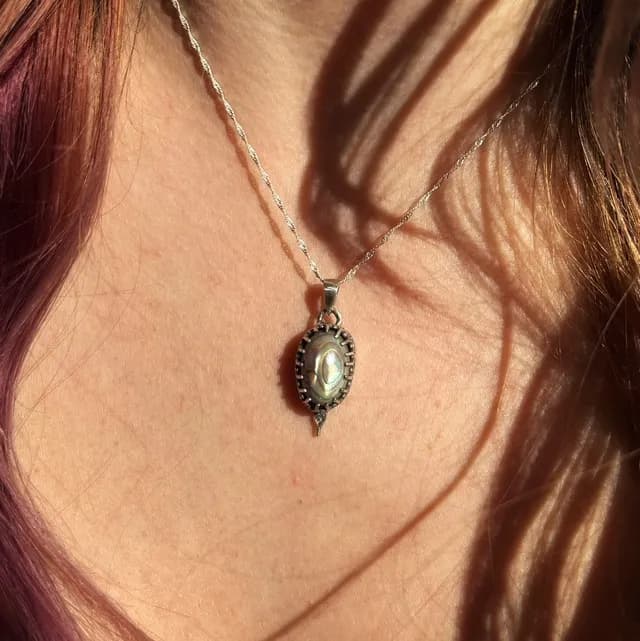Pendentif ovale en argent et nacre abalone aux reflets verts, porté sur le décolleté sous le soleil.