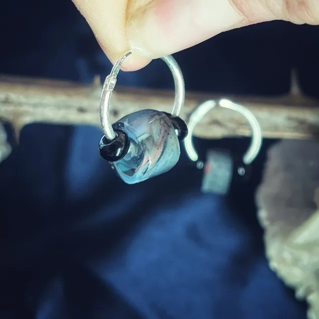 Créole en argent tenue entre les doigts, mettant en valeur la perle de verre aux reflets bleus et gris.