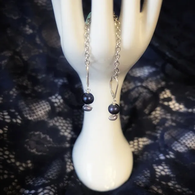 Boucles d'oreilles à perles sombres sur un présentoir main blanc aux ongles rouges, fond dentelle noire.