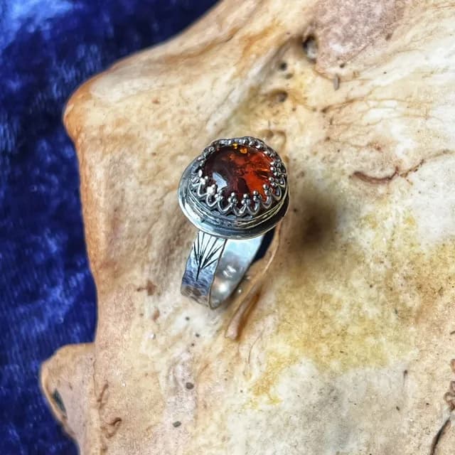 Bague en argent martelé avec une pierre d'ambre ronde et orangée, posée sur un support beige.
