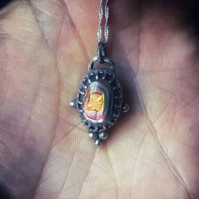 Pendentif ovale en argent patiné avec un cabochon aux reflets orange et rose, posé au creux d'une main.