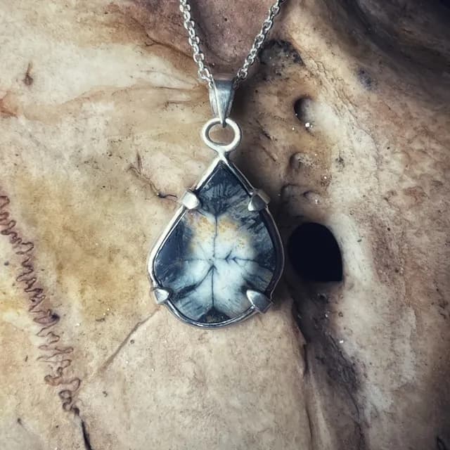 Pendentif en forme de goutte avec une pierre andalousite grise et blanche, posé sur un support texturé.