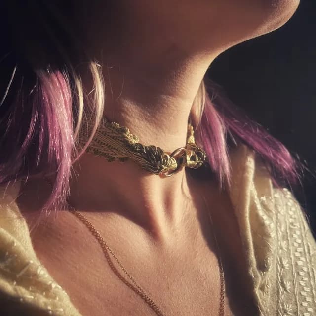Collier ras de cou en galon doré avec deux têtes de lion en laiton reliées par un anneau, porté de face.