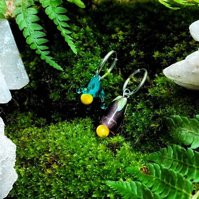 Boucles d'oreilles créoles avec perles en verre bleue et violette sur un lit de mousse verte.