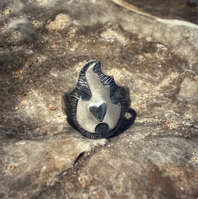 Bague en argent en forme de flamme avec un cœur au centre, posée de face sur une roche grise.