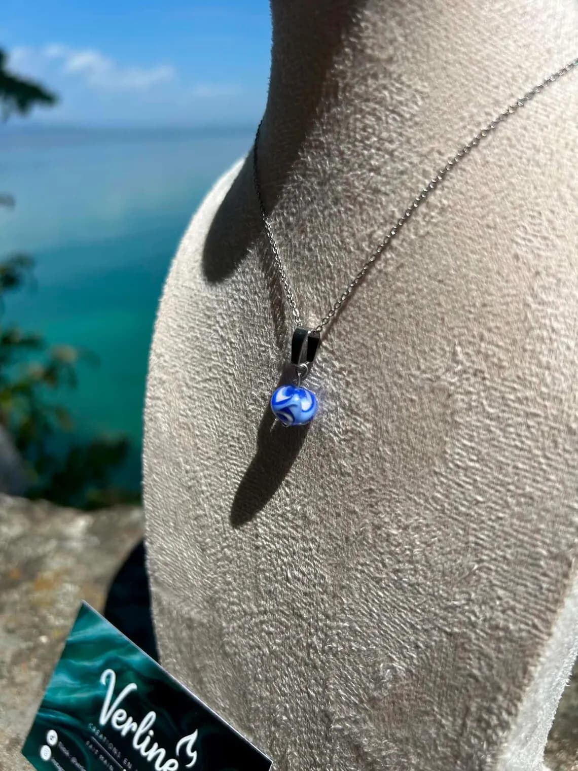 Collier argenté à perle en verre filé bleu et blanc sur un buste de présentation, face à la mer.