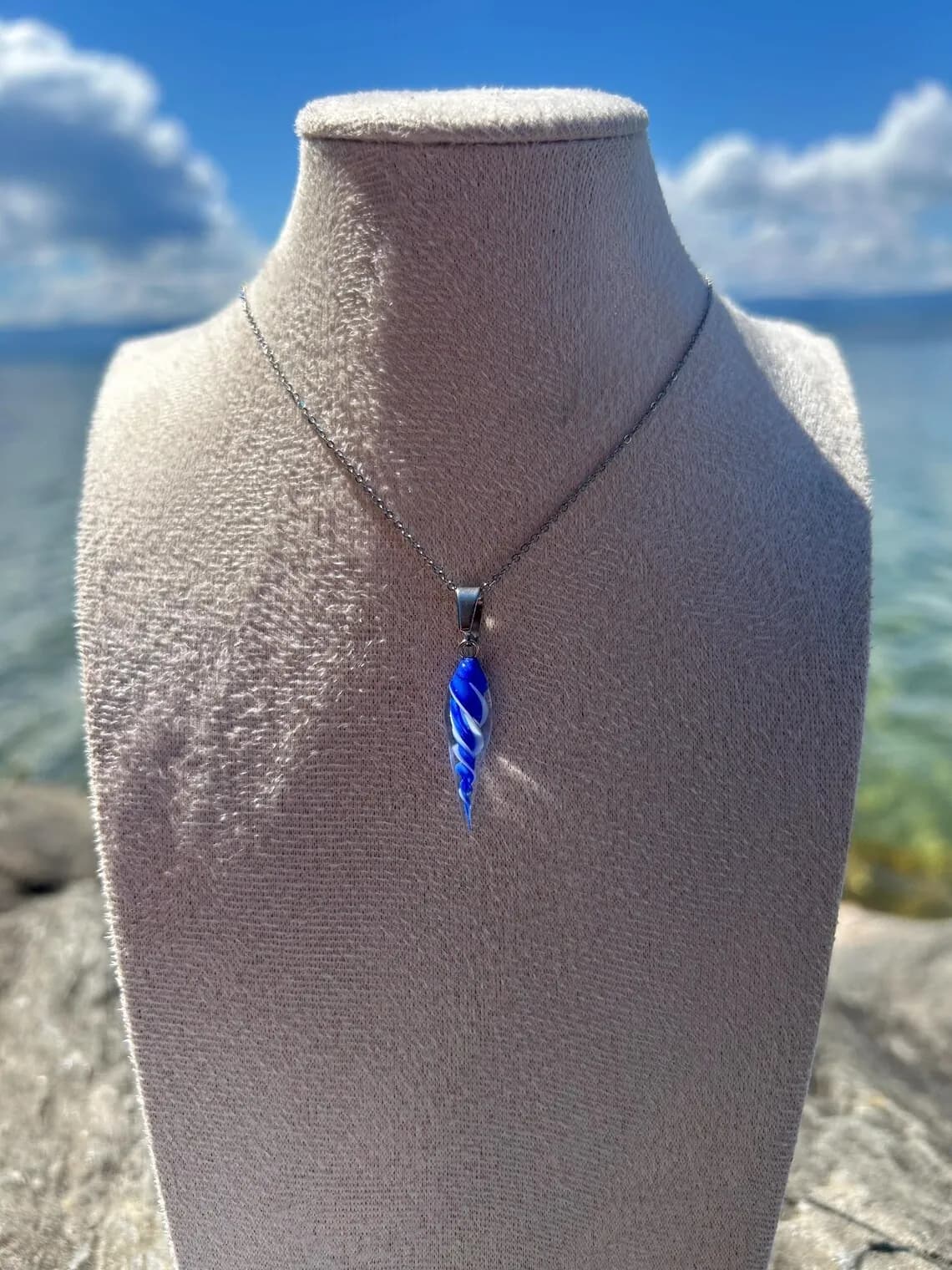 Collier avec pendentif en verre torsadé bleu et blanc sur un buste, devant la mer ensoleillée.