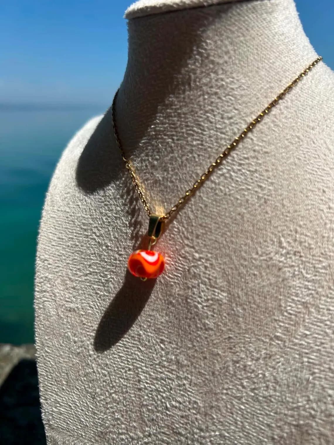 Collier à perle orange et blanche sur un buste beige, devant une mer bleue et une carte de visite.