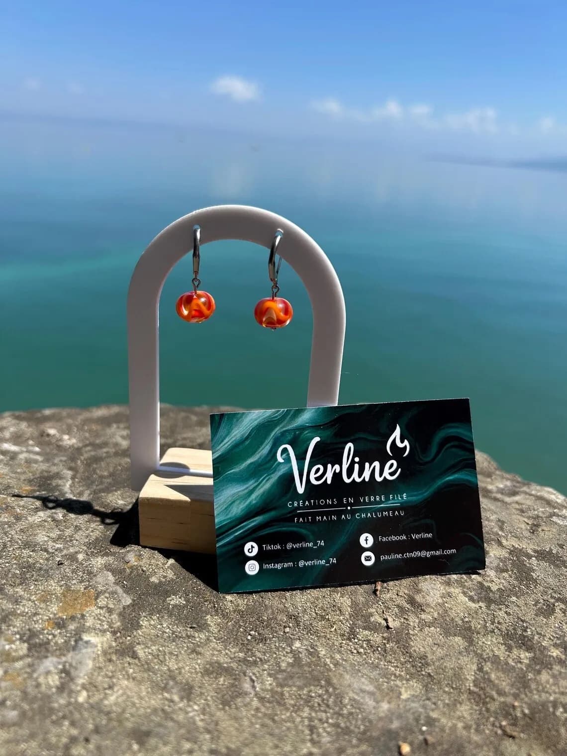 Boucles d'oreilles en verre orange et ambré sur un support blanc, avec la mer bleue en arrière-plan.
