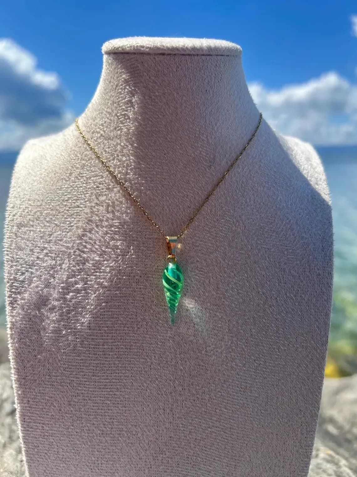 Collier avec pendentif coquillage en verre vert torsadé sur un buste blanc, fond de mer et ciel bleu.