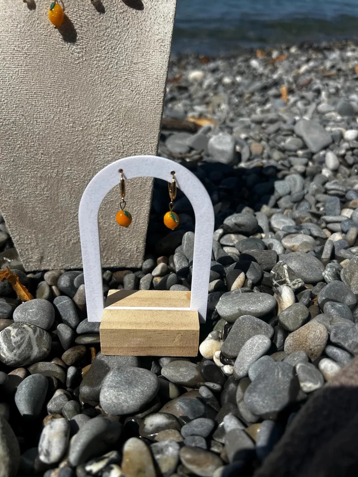 Boucles d'oreilles en forme de petits citrons jaunes suspendues sur un support blanc posé sur des galets.