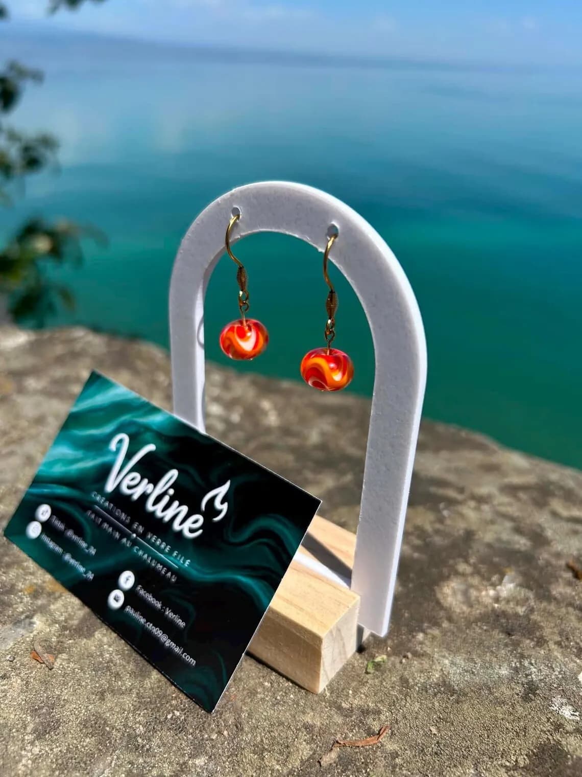 Boucles d'oreilles rondes orange et dorées sur un présentoir blanc, avec la mer bleue en arrière-plan.