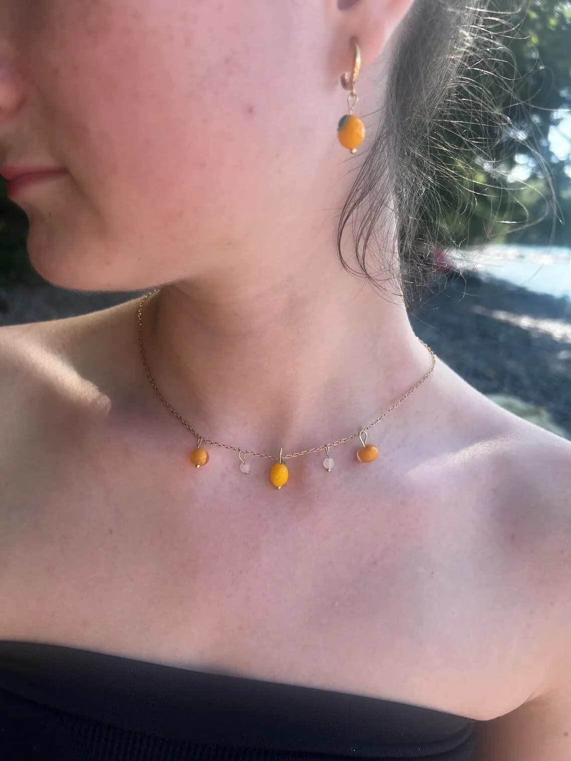 Collier aux pendentifs citrons jaunes sur un présentoir beige, posé sur une plage de galets face à la mer.