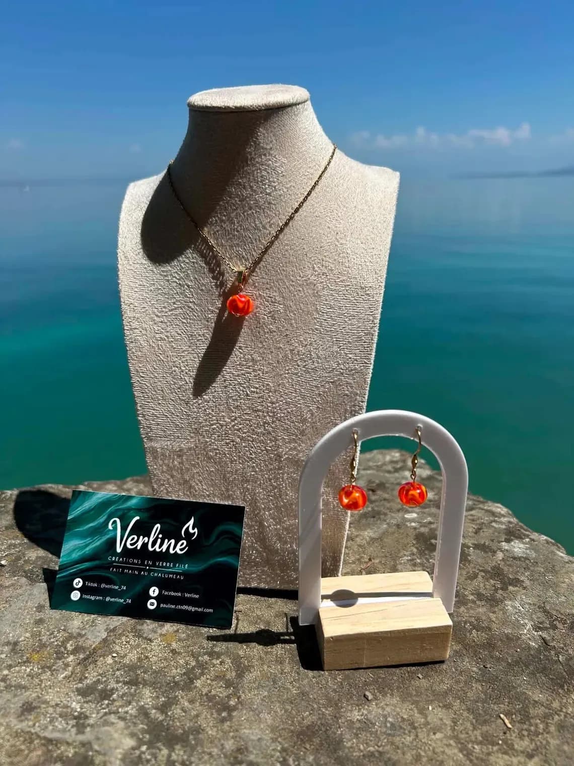 Collier et boucles d'oreilles orange et doré présentés sur un buste et un support devant la mer.