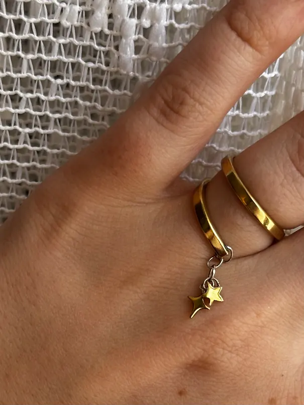 Bague double anneau dorée avec deux petites étoiles suspendues, portée sur un doigt devant un tissu blanc.